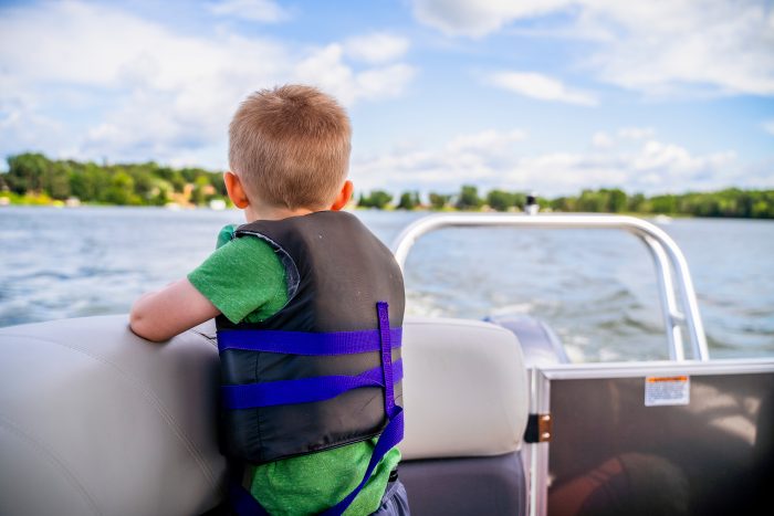 Toddler Boy In Life Vest Looking Out At Lake On Pontoon Boat